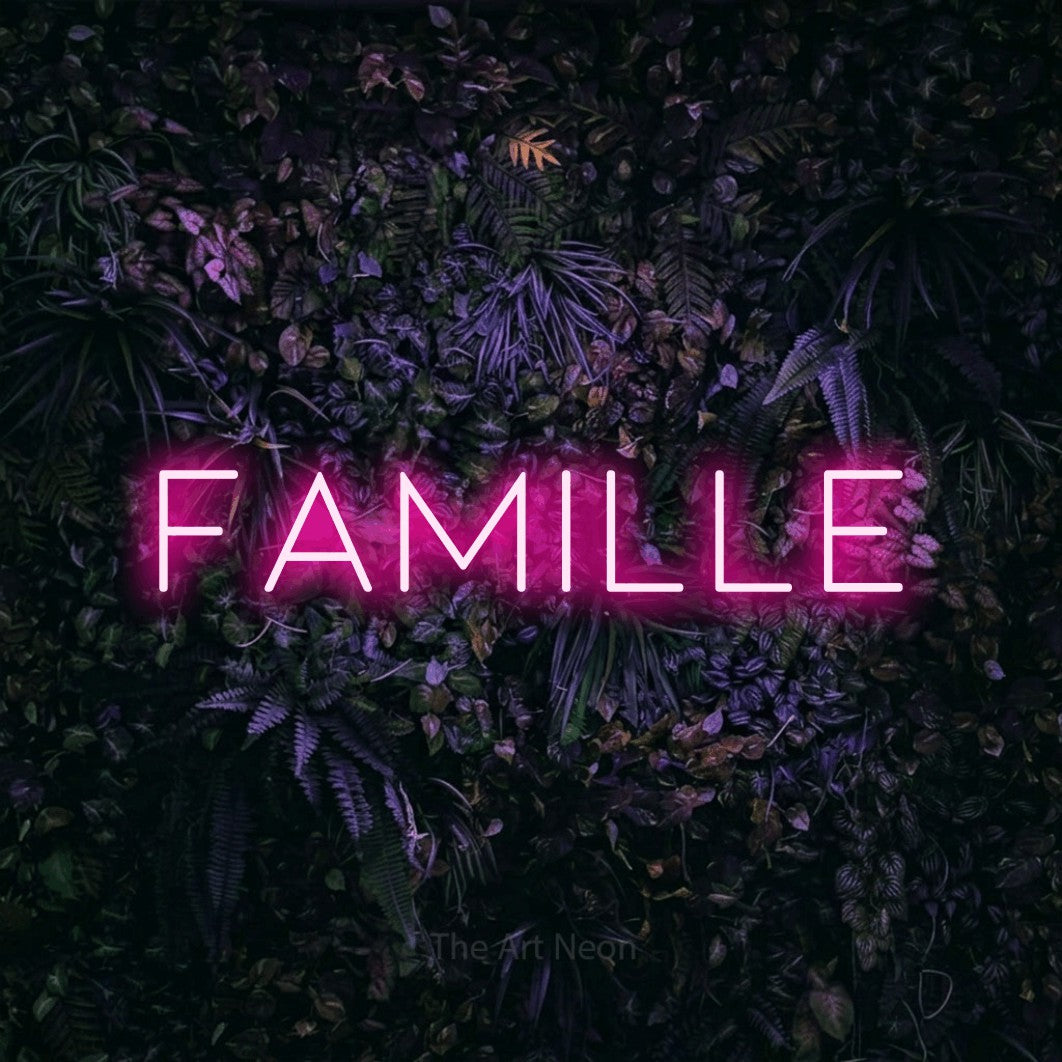 “Famille” LED neon sign in hot pink, with thin, widely spaced lettering, shown against a dark wall with purple flowers.