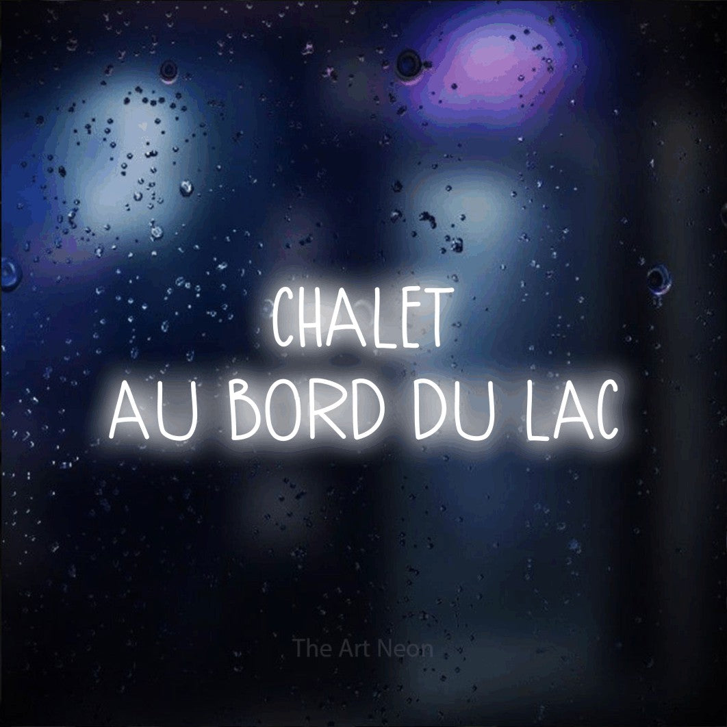 “Chalet au bord du lac” LED neon sign in cool white, with narrow hand-drawn lettering across 2 lines, shown against glass with blue and purple reflections.