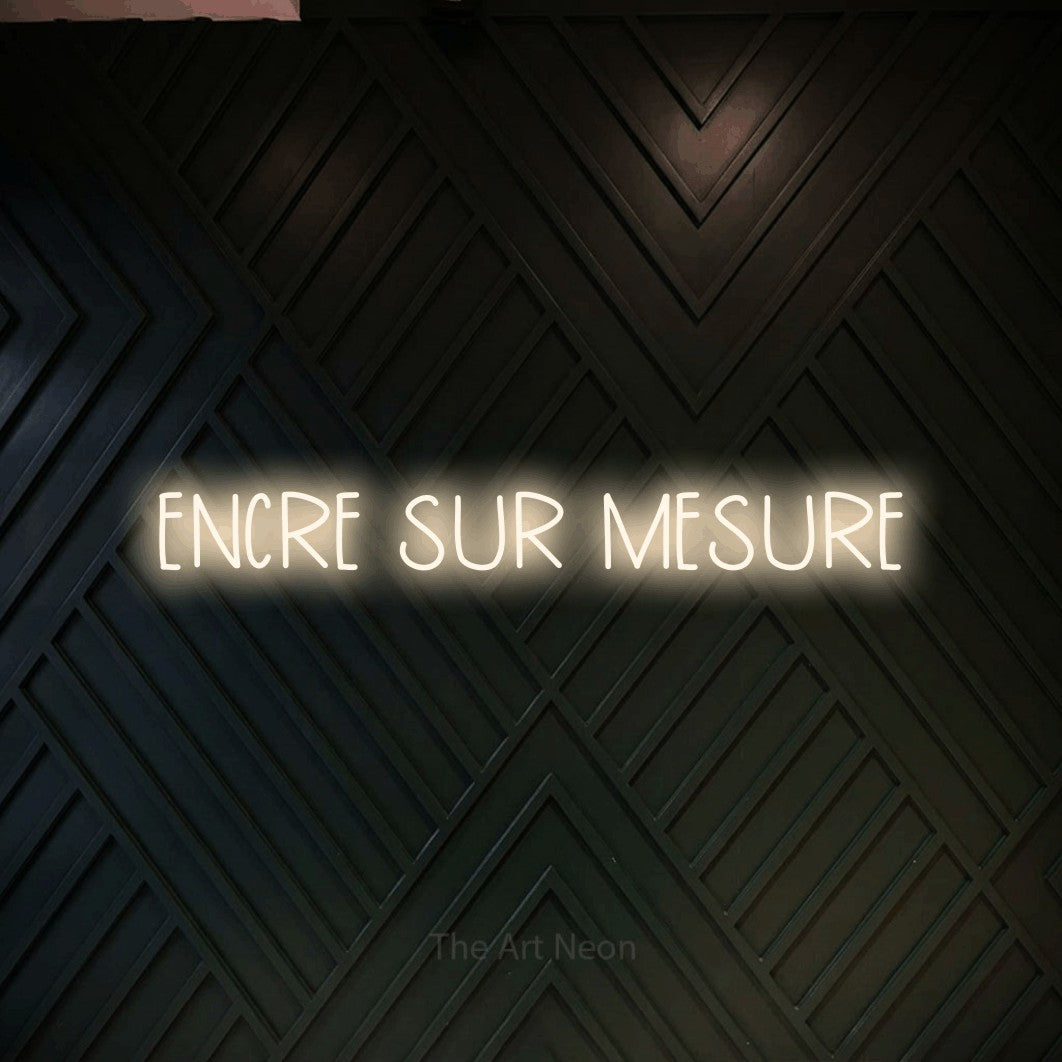 “Encre sur mesure” LED neon sign in warm white, with narrow hand-drawn lettering, shown against a black chevron-patterned wood wall.