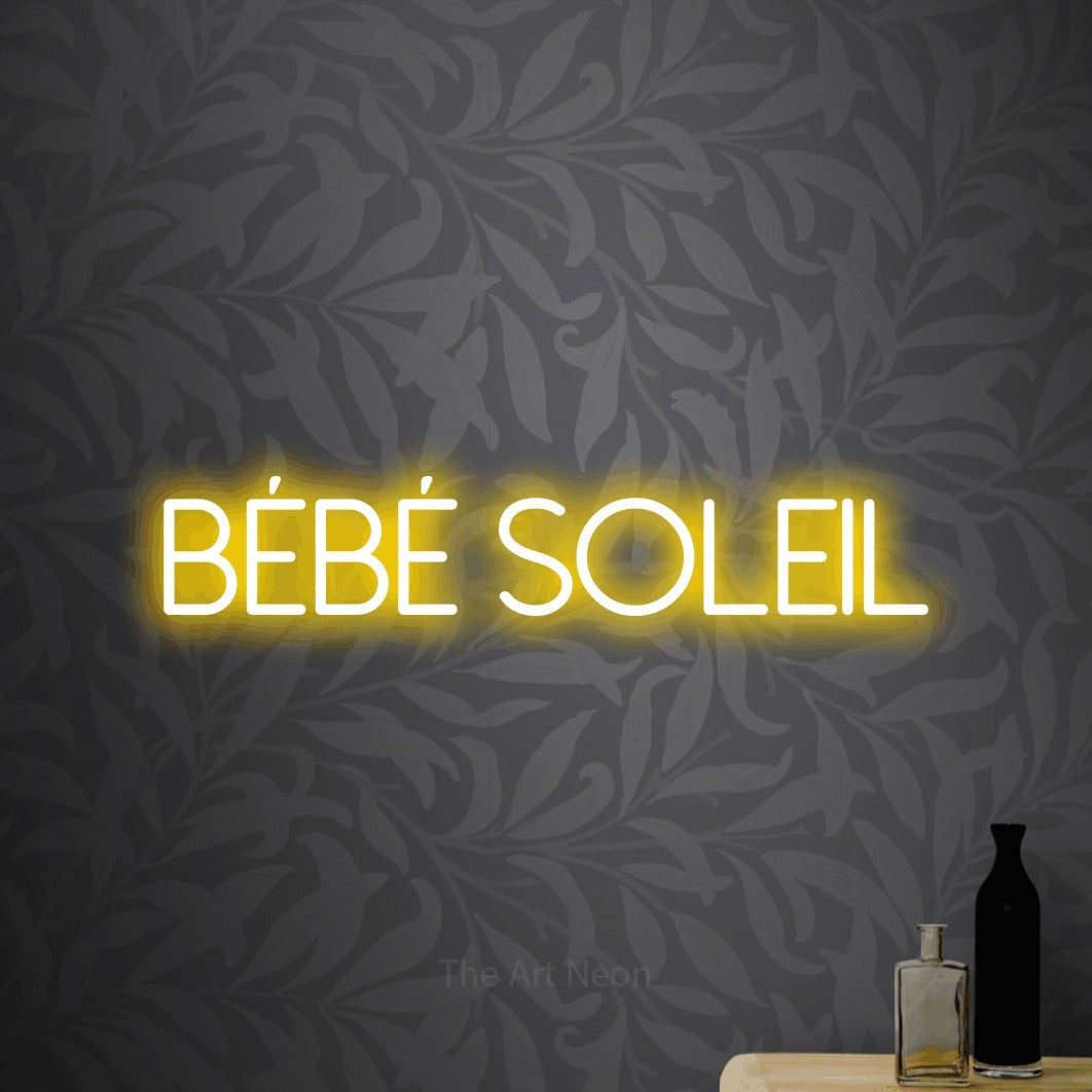 Décoration murale en néon LED « Bébé soleil » en jaune, en lettres fines sans empattement, installée sur un papier peint botanique gris, pour une fête prénatale.