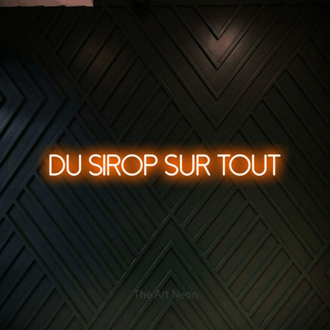 “Du sirop sur tout” LED neon sign in orange, with thin sans-serif lettering, shown against a black chevron-patterned wood wall.