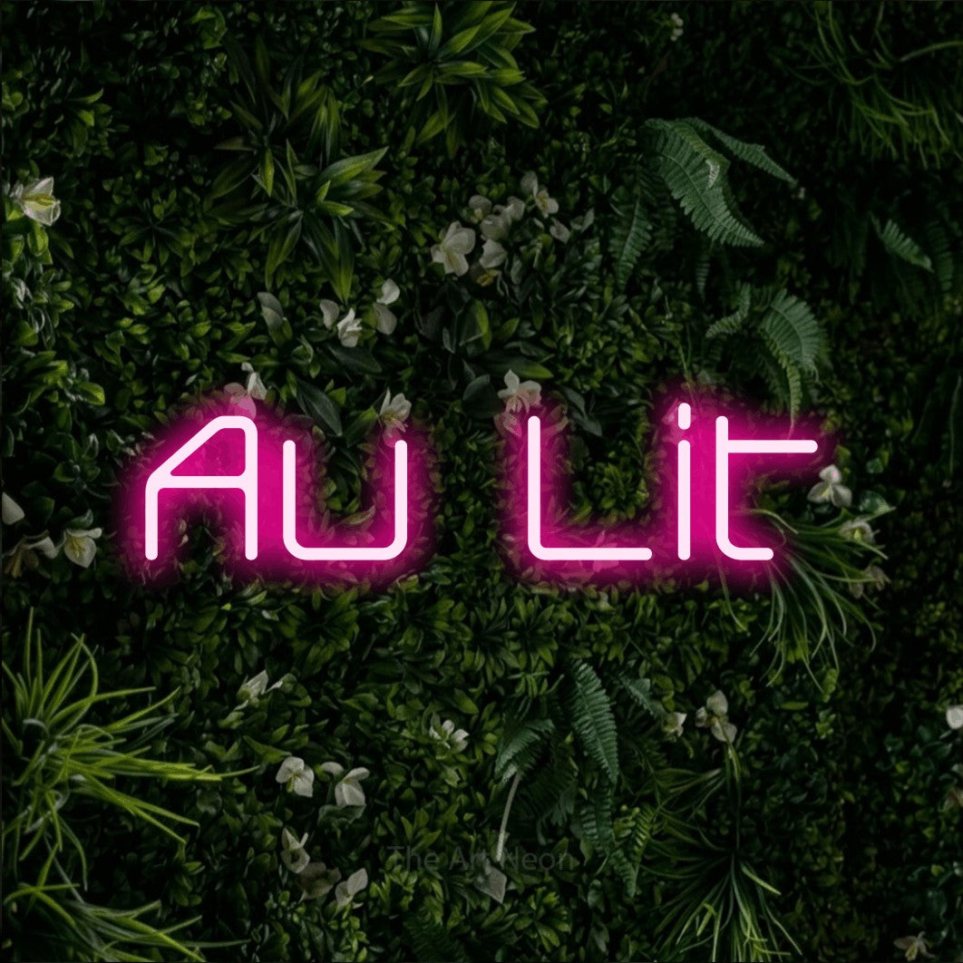 “Au lit” LED neon sign in hot pink, with rounded geometric lettering, shown against a dense green plant wall.