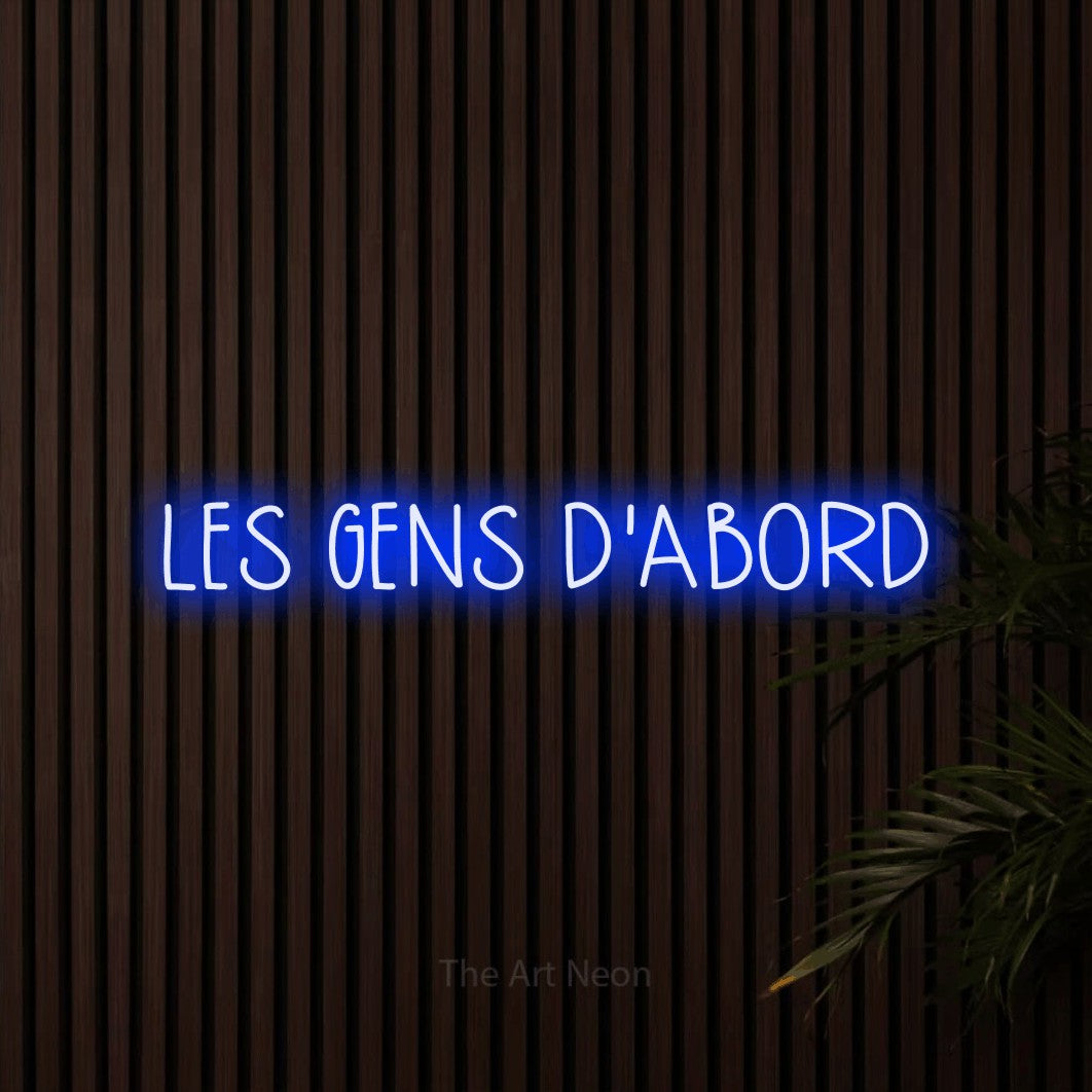 “Les gens d'abord” LED neon sign in blue, with narrow hand-drawn lettering, shown against dark wooden wall slats.
