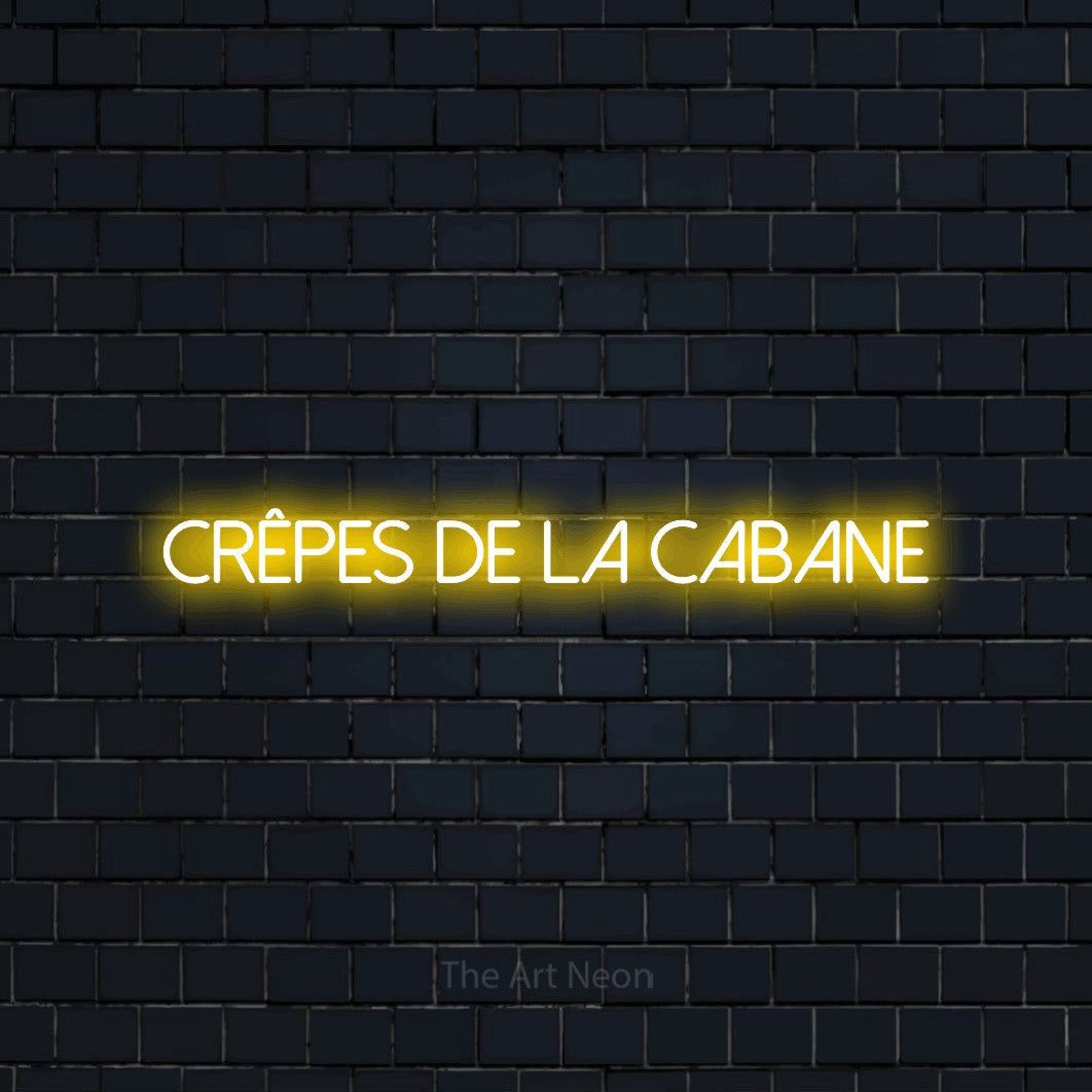 “Crêpes de la cabane” LED neon sign in yellow, with thin sans-serif lettering, shown against a dark navy brick wall.