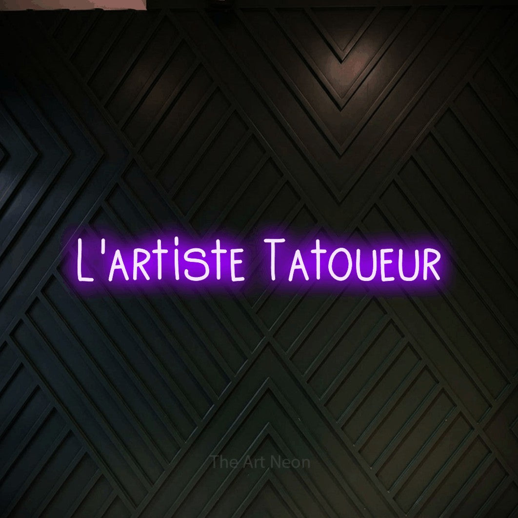 “L'artiste tatoueur” LED neon sign in purple, with narrow hand-drawn lettering, shown against a black chevron-patterned wood wall.