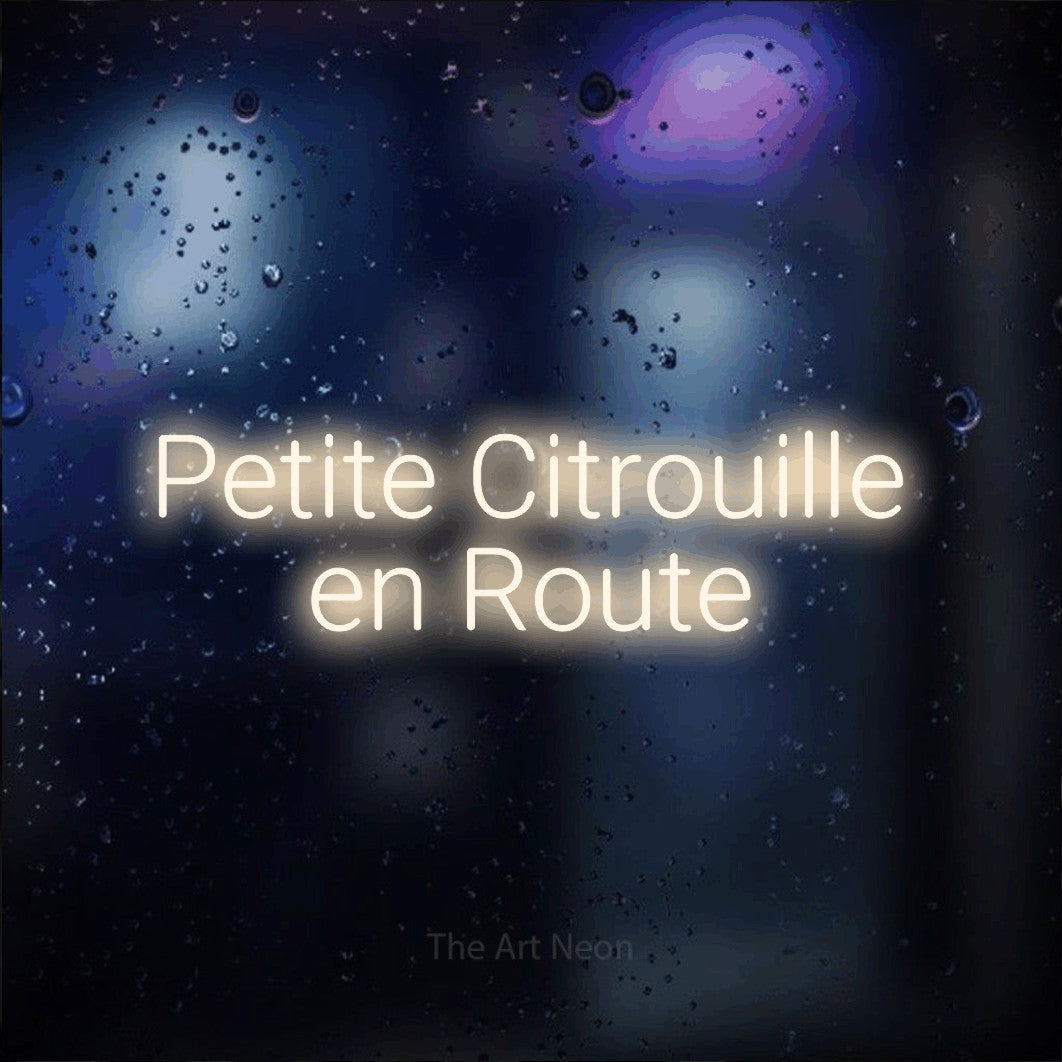 “Petite citrouille en route” LED neon sign in warm white, with rounded sans-serif lettering across 2 lines, shown against glass with blue and purple reflections.