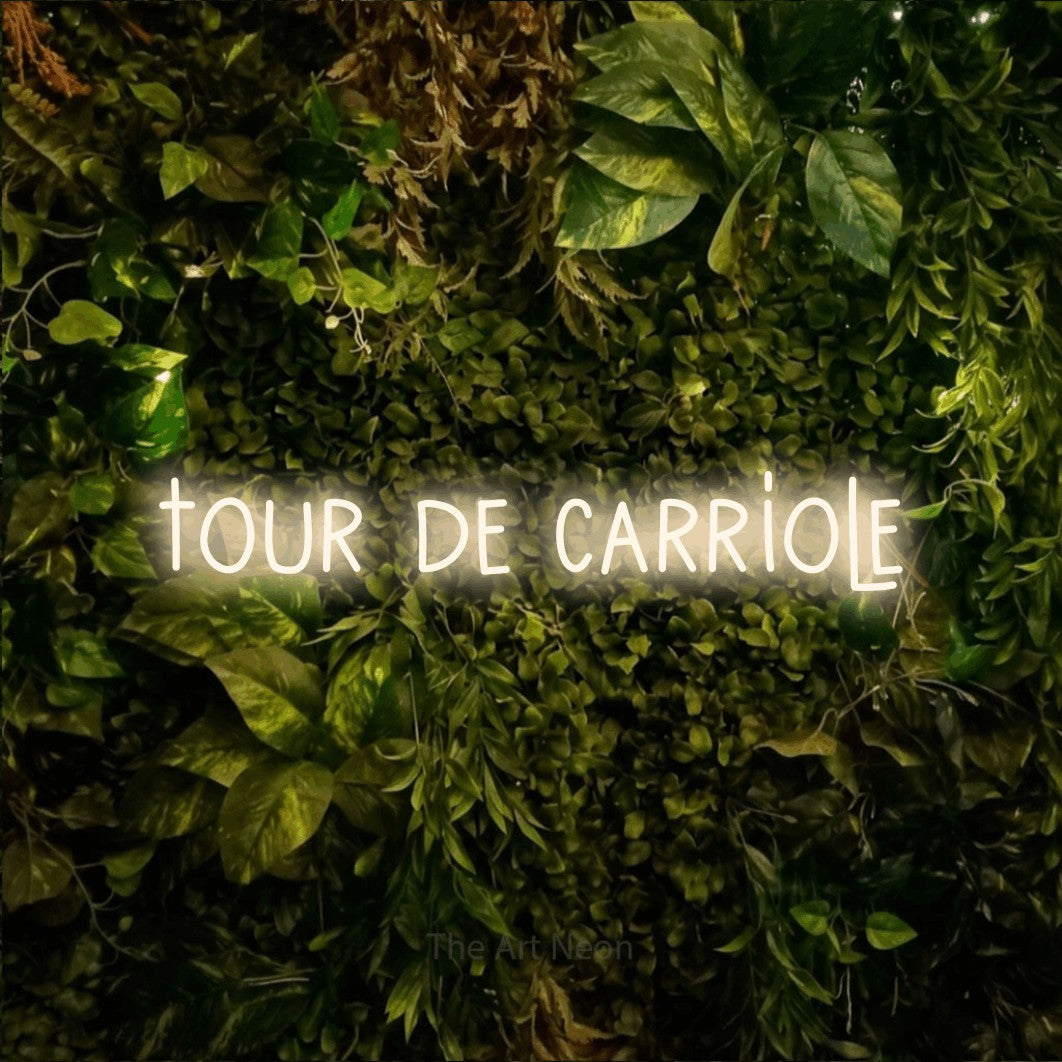 “Tour de carriole” LED neon sign in warm white, with narrow hand-drawn lettering, shown against bright green and yellow foliage.