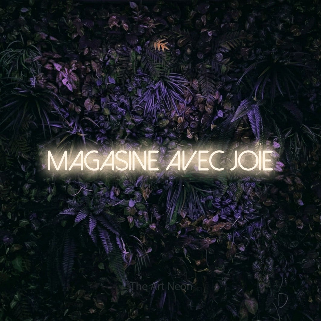 “Magasine avec joie” LED neon sign in warm white, with thin sans-serif lettering, shown against a dark wall with purple flowers.