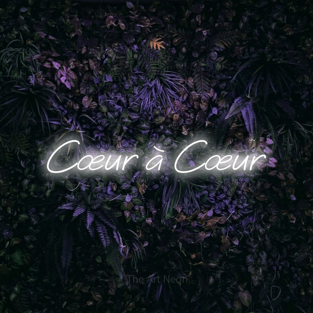 “Cœur à cœur” LED neon sign in cool white, with slanted cursive lettering, shown against a dark wall with purple flowers.