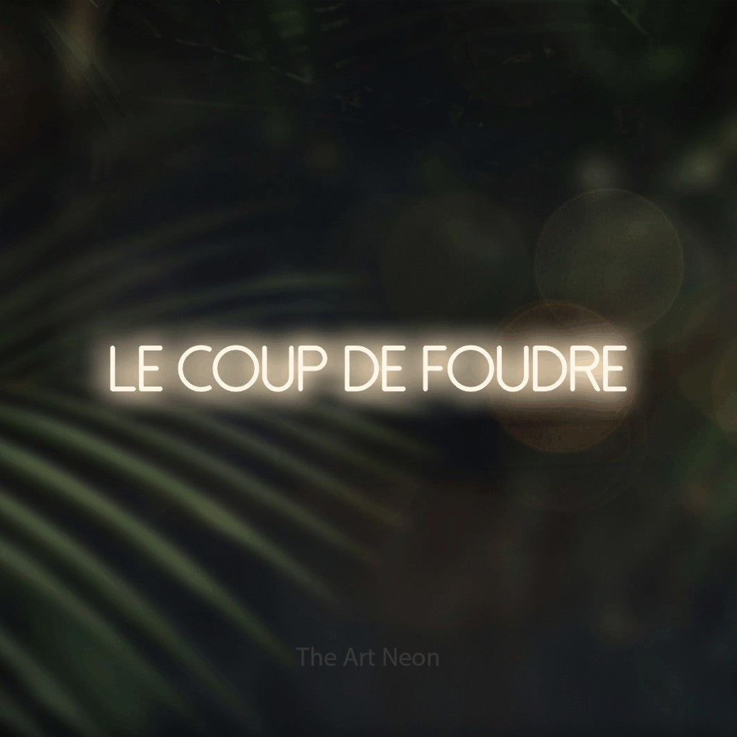 “Le coup de foudre” LED neon sign in warm white, with thin sans-serif lettering, shown against a blurred palm and bokeh-light backdrop.
