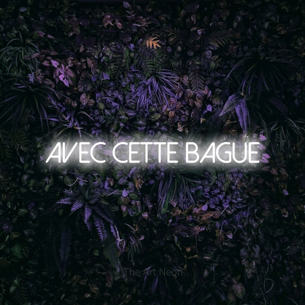 Aperçu d’une enseigne néon LED « Avec cette bague » en blanc froid, en lettres fines sans empattement, photographiée sur un mur sombre aux fleurs violettes, pour une réception de mariage.