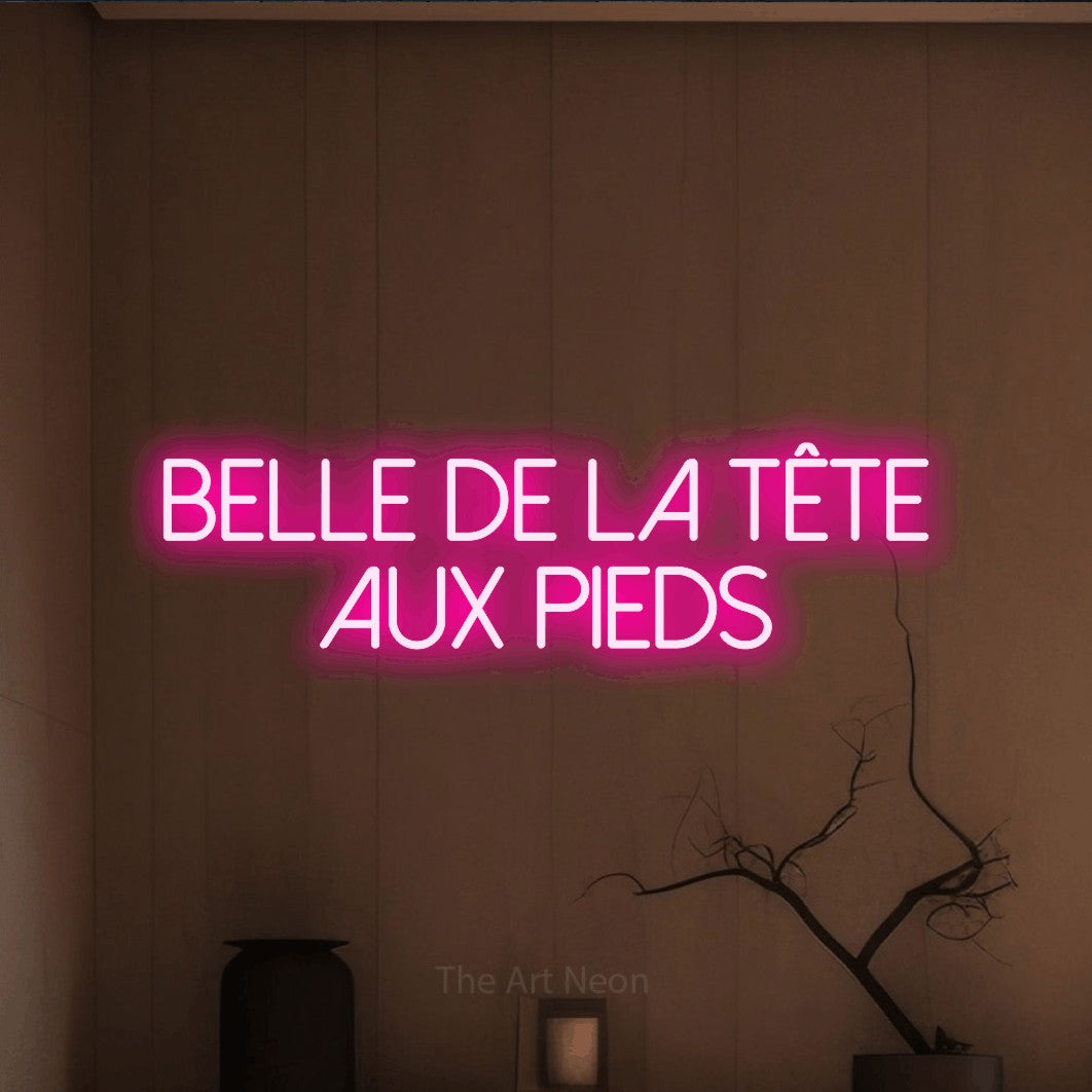 “Belle de la tête aux pieds” LED neon sign in hot pink, with thin sans-serif lettering across 2 lines, shown against a simple earth-toned interior.