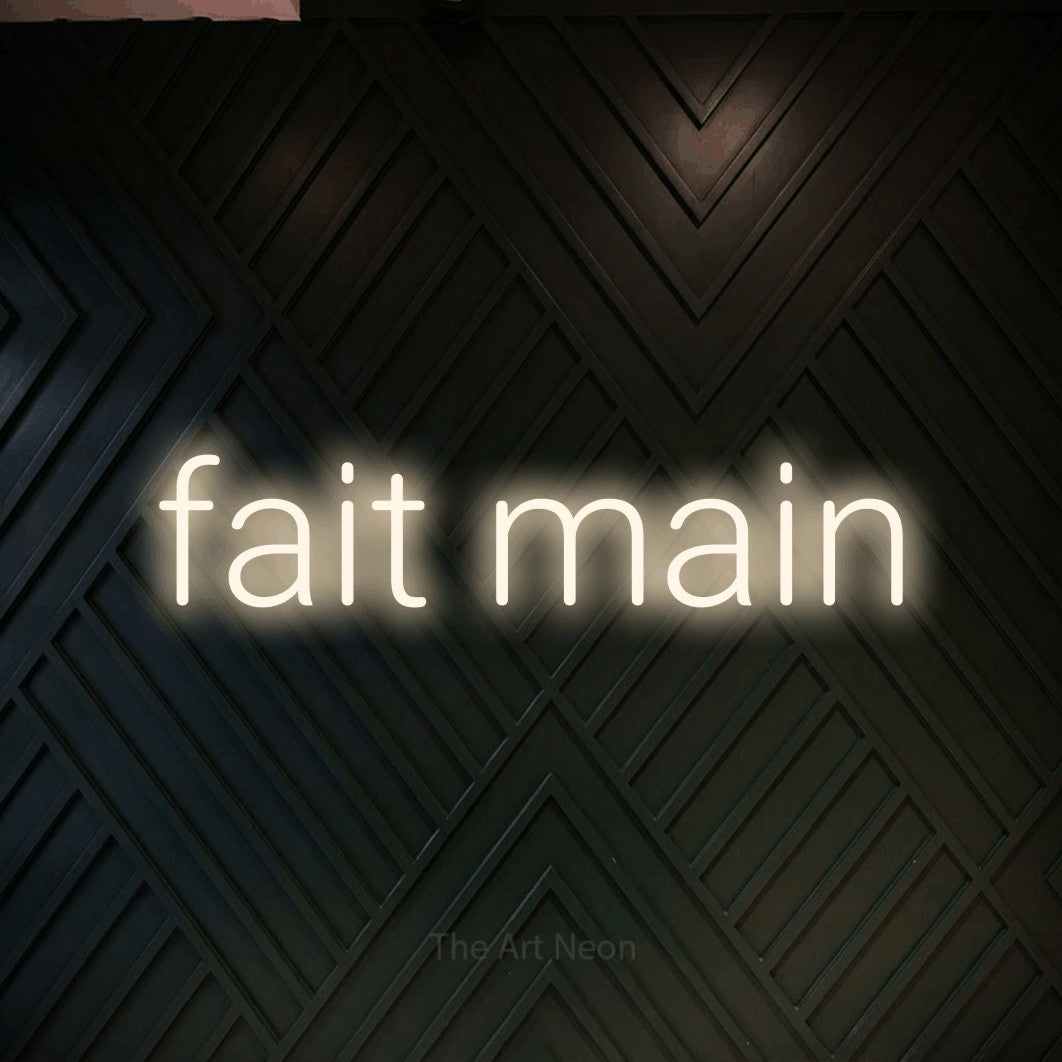 “Fait main” LED neon sign in warm white, with rounded sans-serif lettering, shown against a black chevron-patterned wood wall.