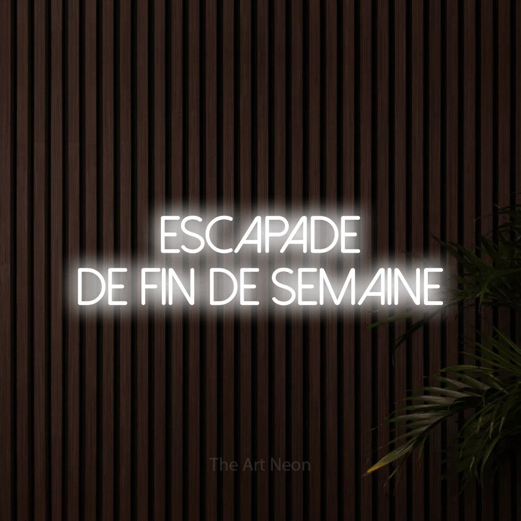 “Escapade de fin de semaine” LED neon sign in cool white, with thin sans-serif lettering across 2 lines, shown against dark wooden wall slats.