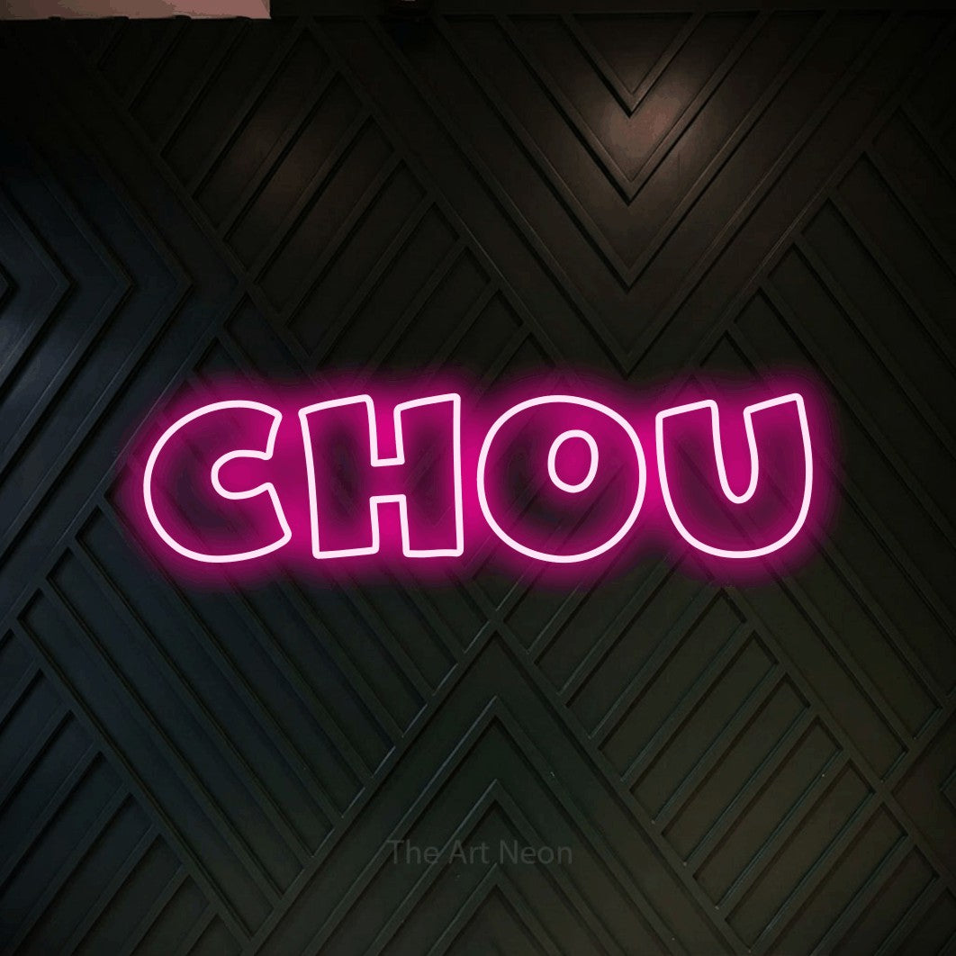“Chou” LED neon sign in hot pink, with wide lettering with irregular outlines, shown against a black chevron-patterned wood wall.