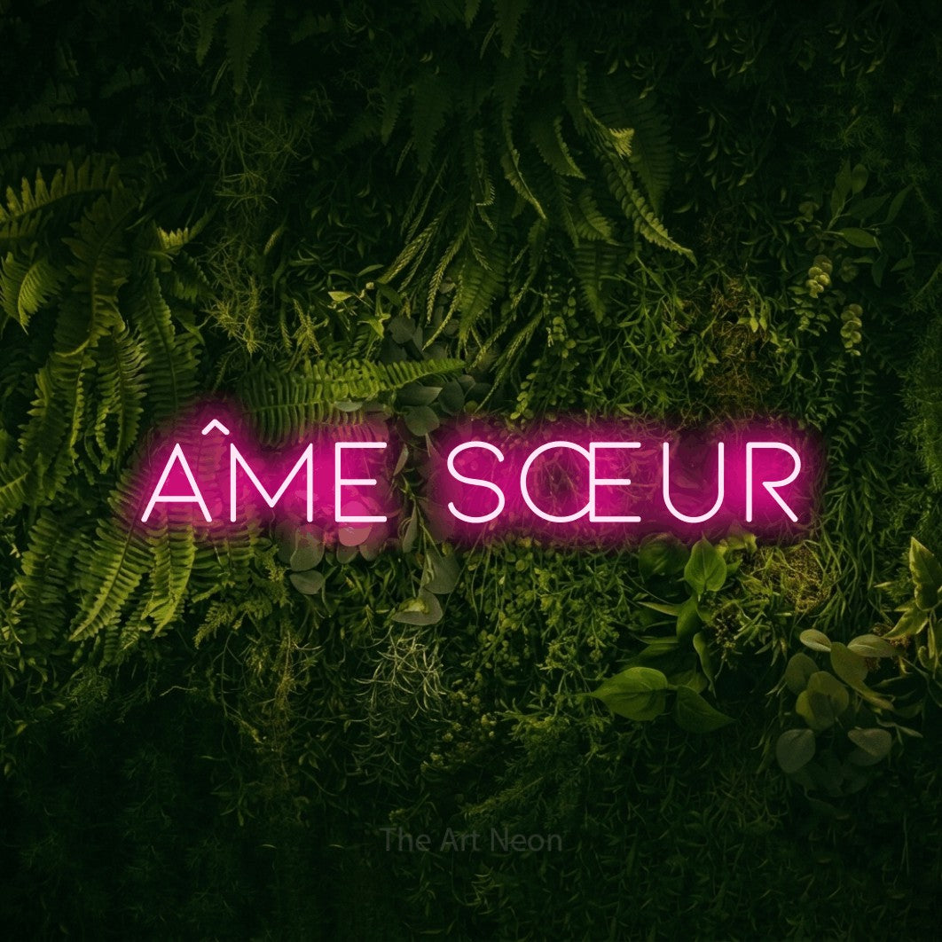 “Âme sœur” LED neon sign in hot pink, with thin, widely spaced lettering, shown against a dark moss and forest backdrop.