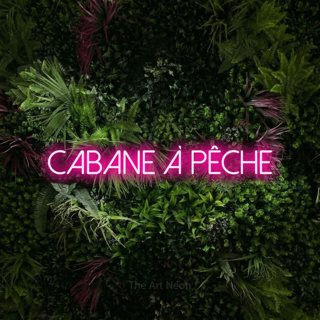 “Cabane à pêche” LED neon sign in hot pink, with thin sans-serif lettering, shown against a plant wall with pink flowers.