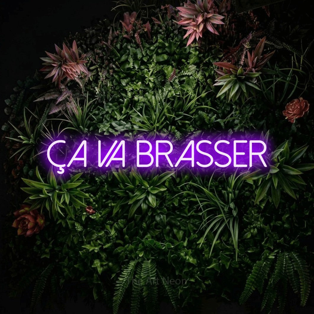 “Ça va brasser” LED neon sign in purple, with thin sans-serif lettering, shown against a flowering plant wall.