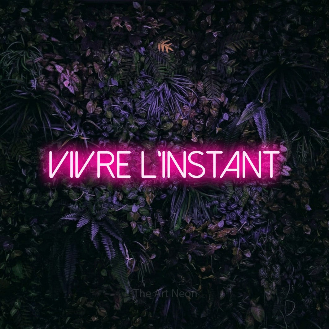 “Vivre l'instant” LED neon sign in hot pink, with thin sans-serif lettering, shown against a dark wall with purple flowers.