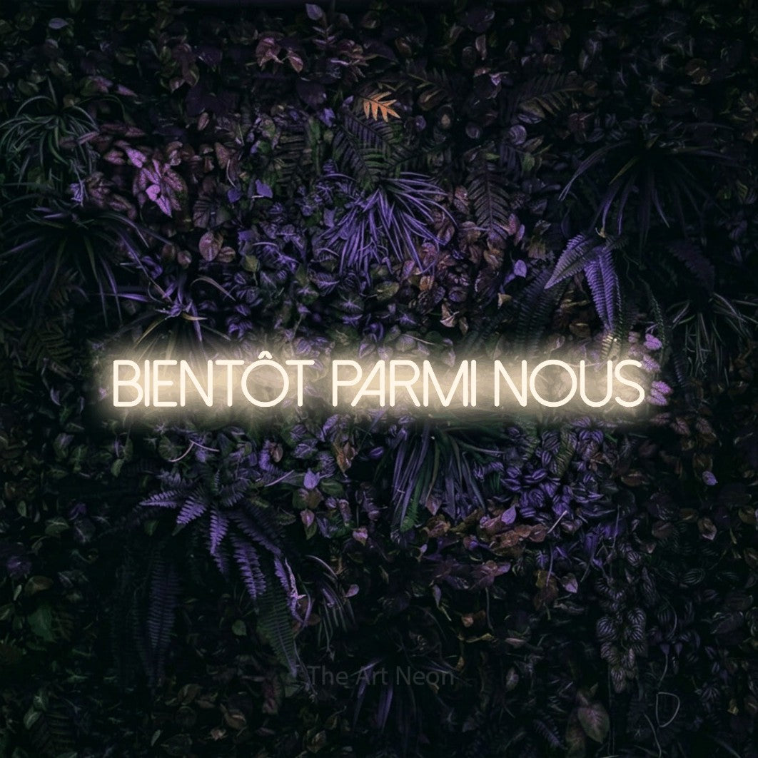 “Bientôt parmi nous” LED neon sign in warm white, with thin sans-serif lettering, shown against a dark wall with purple flowers.