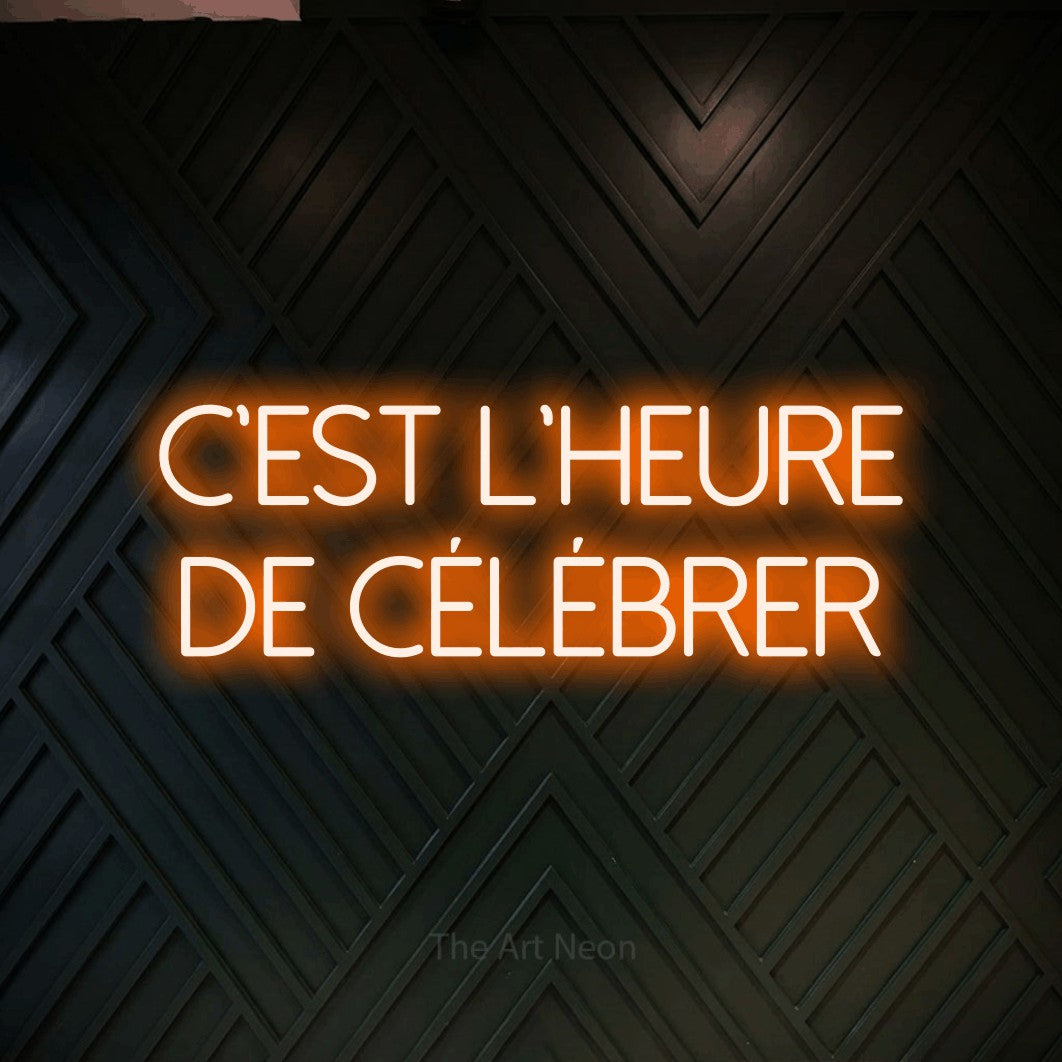 “C'est l'heure de célébrer” LED neon sign in orange, with thin sans-serif lettering across 2 lines, shown against a black chevron-patterned wood wall.