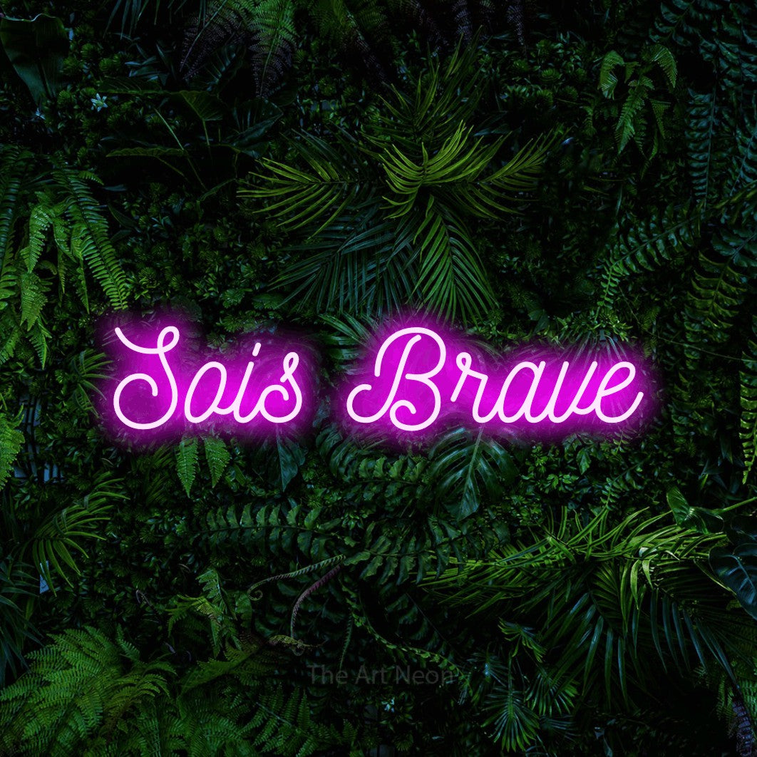 “Sois brave” LED neon sign in pink, with ornate cursive lettering, shown against dense tropical foliage.