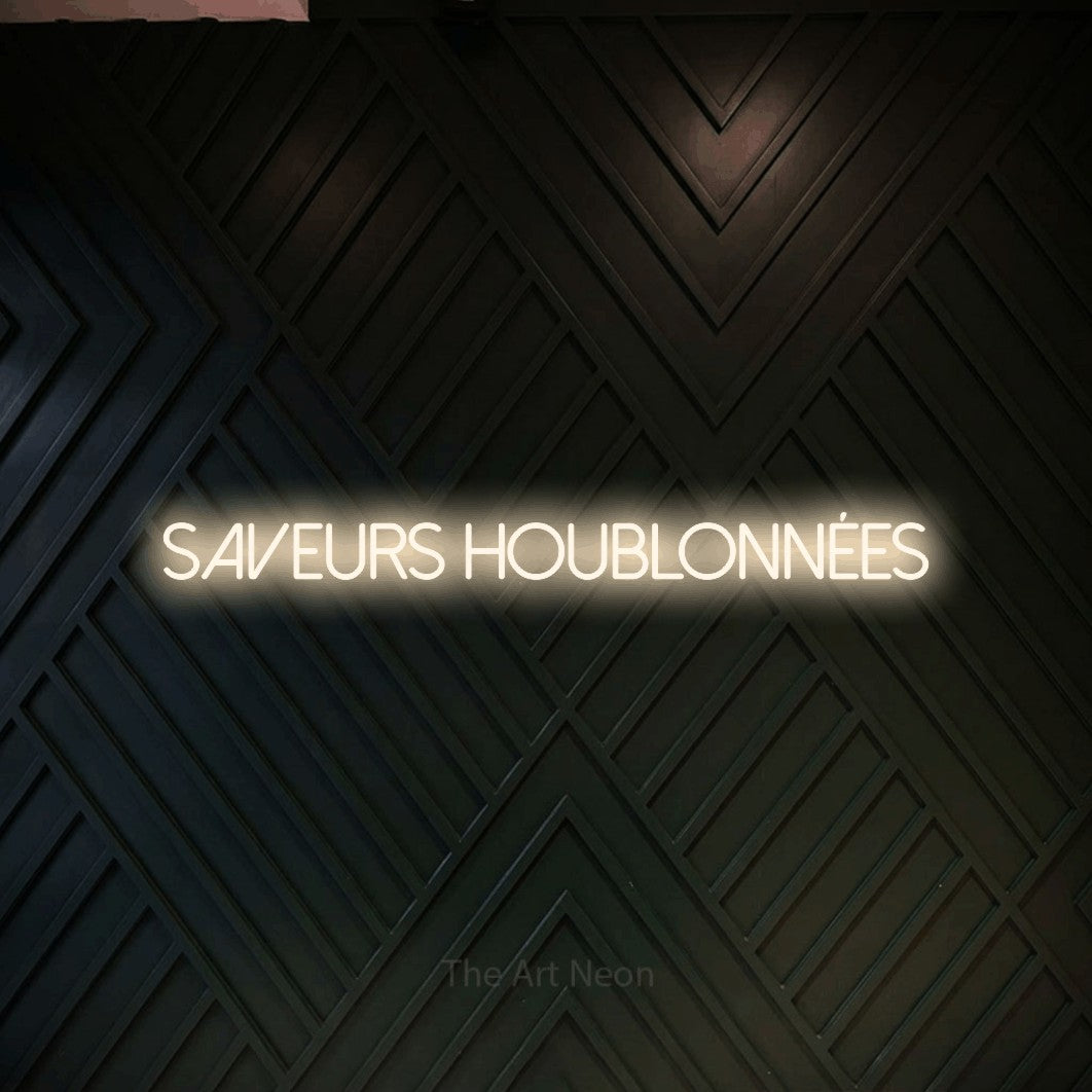 “Saveurs houblonnées” LED neon sign in warm white, with thin sans-serif lettering, shown against a black chevron-patterned wood wall.