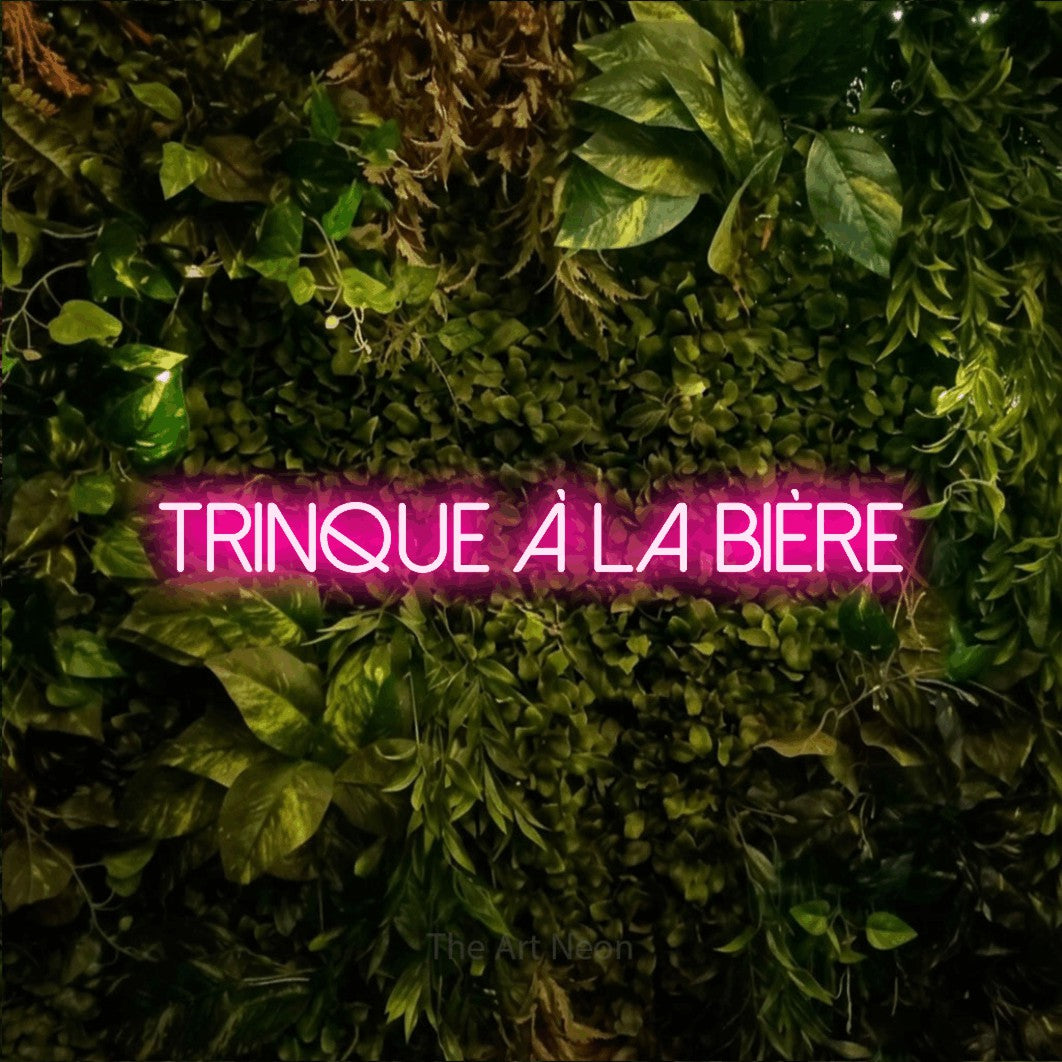 “Trinque à la bière” LED neon sign in hot pink, with thin sans-serif lettering, shown against bright green and yellow foliage.