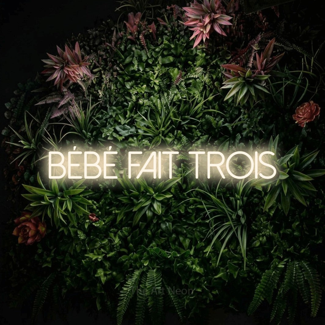 Lettrage en néon LED « Bébé fait trois » en blanc chaud, en lettres fines sans empattement, présenté sur un mur végétal fleuri, pour une fête prénatale.
