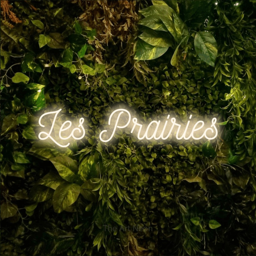 “Les prairies” LED neon sign in warm white, with ornate cursive lettering, shown against bright green and yellow foliage.
