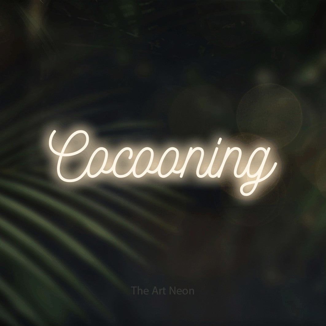 “Cocooning” LED neon sign in warm white, with ornate cursive lettering, shown against a blurred palm and bokeh-light backdrop.