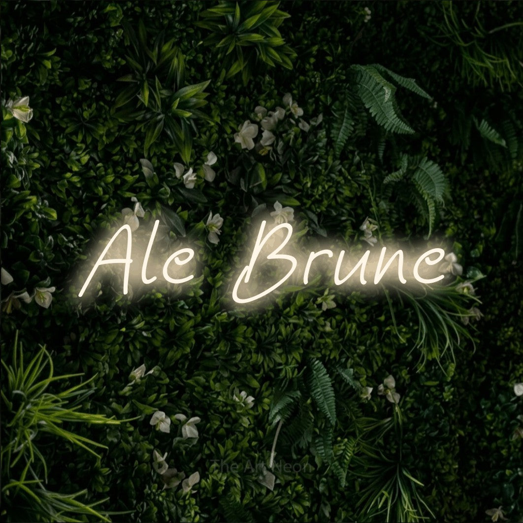 “Ale brune” LED neon sign in warm white, with slanted handwritten lettering, shown against a dense green plant wall.