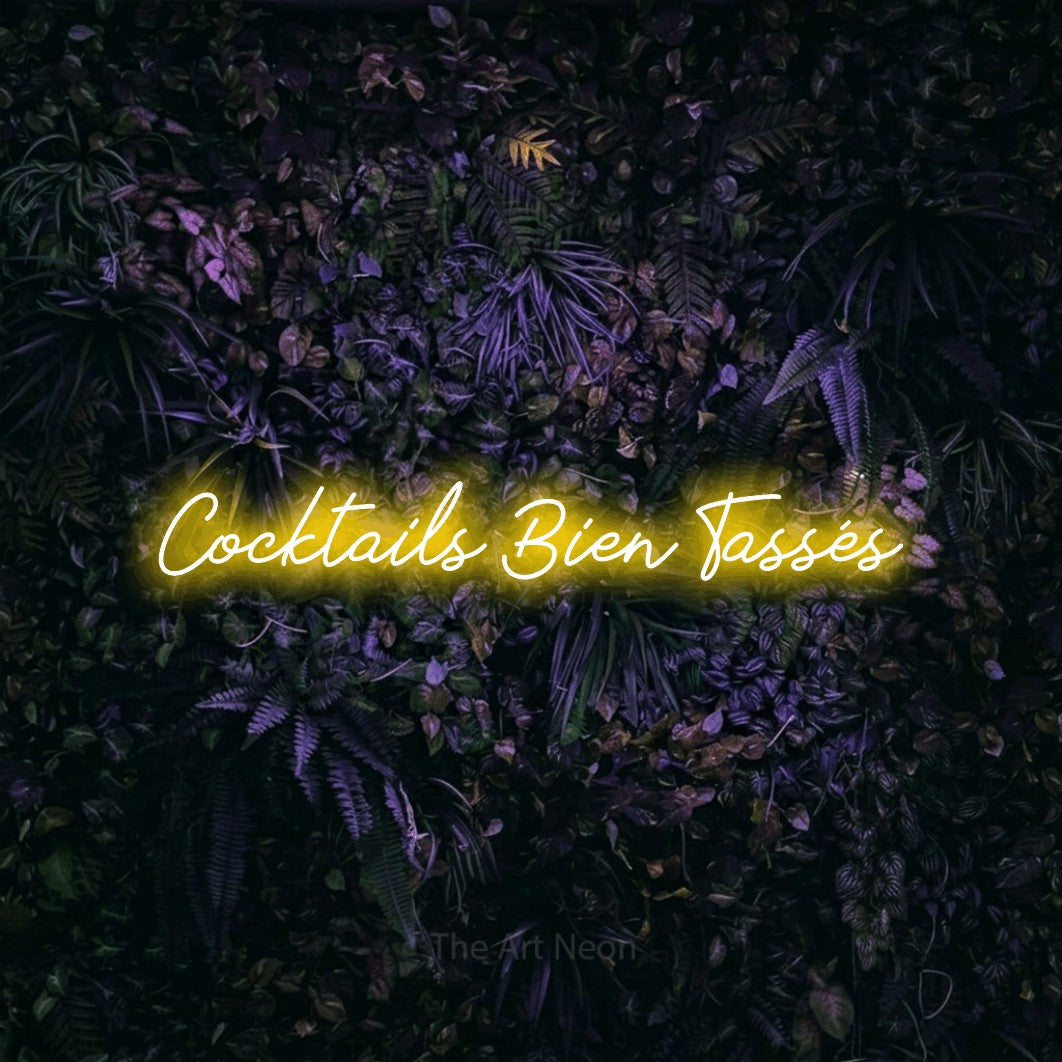 “Cocktails bien tassés” LED neon sign in yellow, with cursive lettering, shown against a dark wall with purple flowers.
