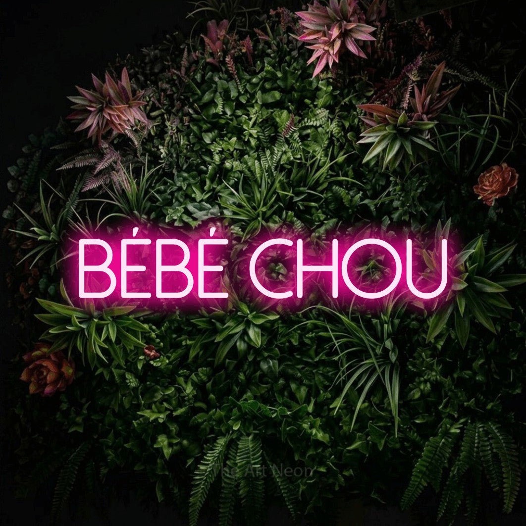 Composition en néon LED « Bébé chou » en rose vif, en lettres fines sans empattement, mise en valeur sur un mur végétal fleuri, pour une fête prénatale.