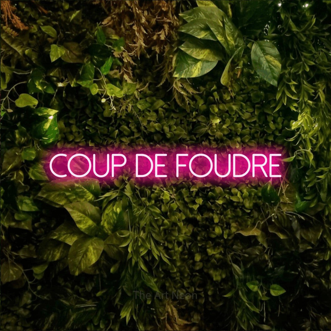 “Coup de foudre” LED neon sign in hot pink, with thin sans-serif lettering, shown against bright green and yellow foliage.