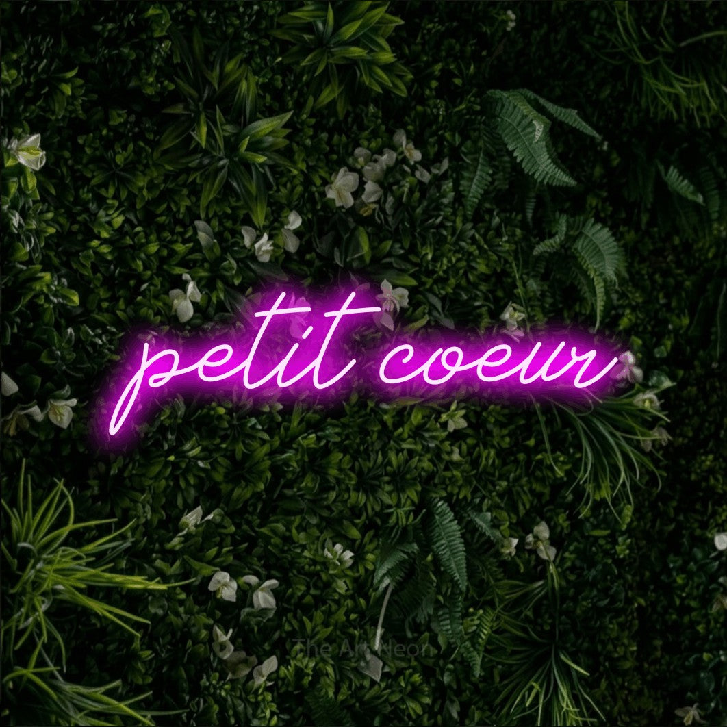 “Petit cœur” LED neon sign in pink, with cursive lettering, shown against a dense green plant wall.