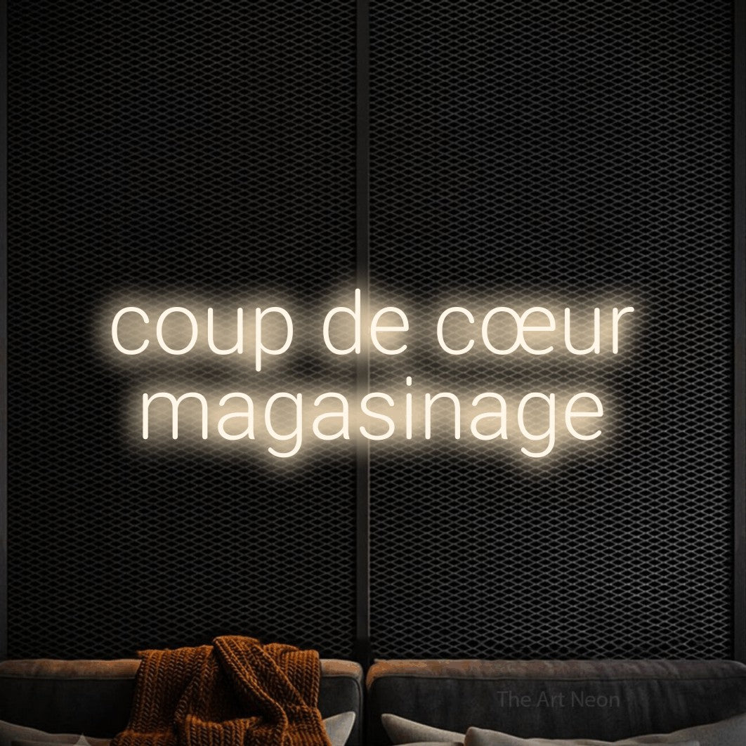 “Coup de cœur magasinage” LED neon sign in warm white, with rounded sans-serif lettering across 2 lines, shown against a modern dark-toned seating area.