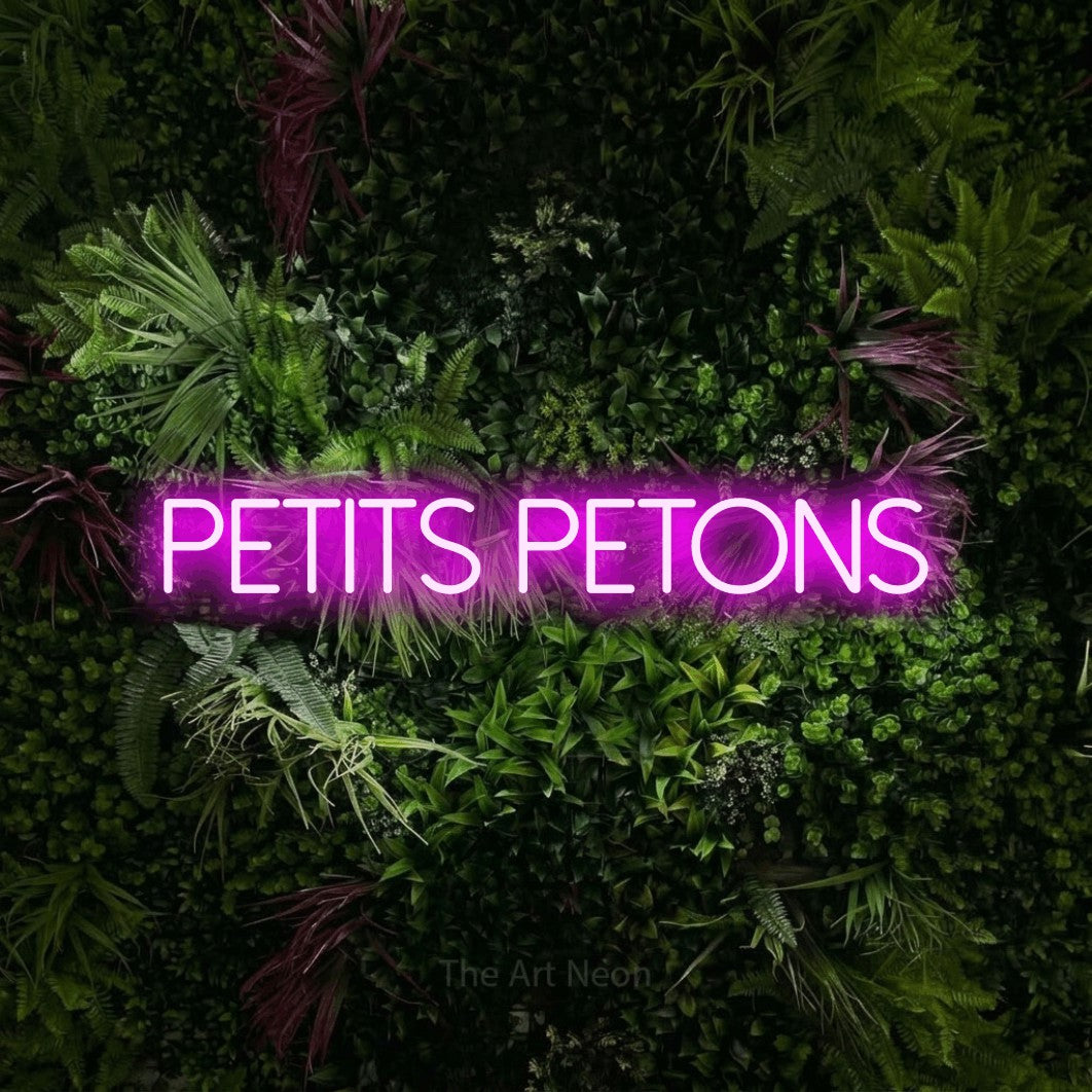 “Petits petons” LED neon sign in pink, with thin sans-serif lettering, shown against a plant wall with pink flowers.