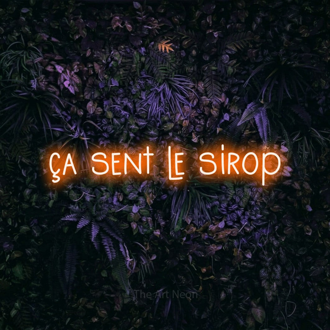 “Ça sent le sirop” LED neon sign in orange, with narrow hand-drawn lettering, shown against a dark wall with purple flowers.