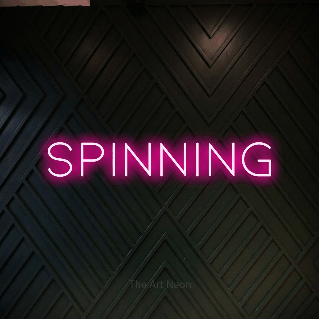 Composition en néon LED « Spinning » en rose vif, en lettres fines et espacées, mise en valeur sur un mur en bois noir à motif chevron, pour un espace d’entraînement.