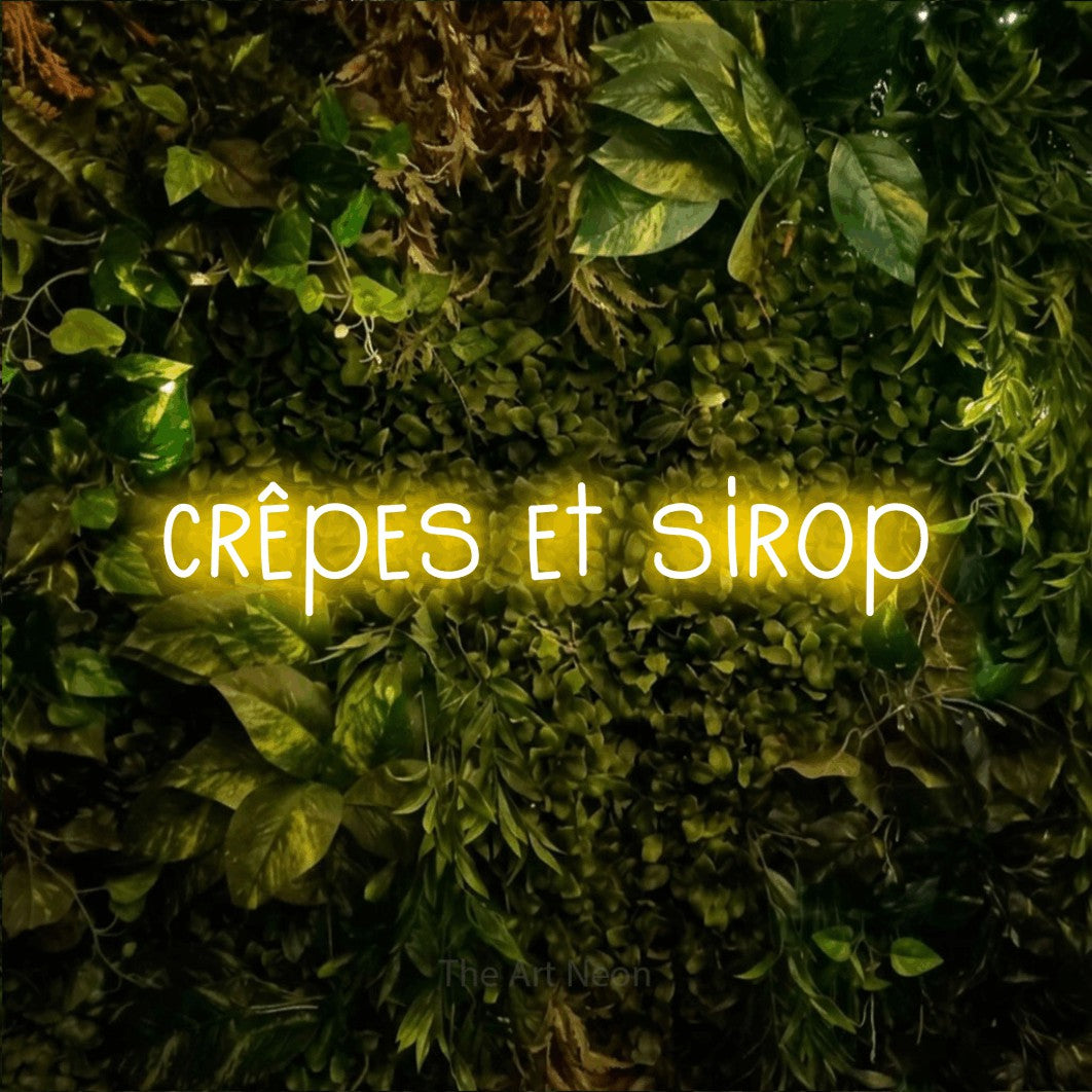 “Crêpes et sirop” LED neon sign in yellow, with narrow hand-drawn lettering, shown against bright green and yellow foliage.
