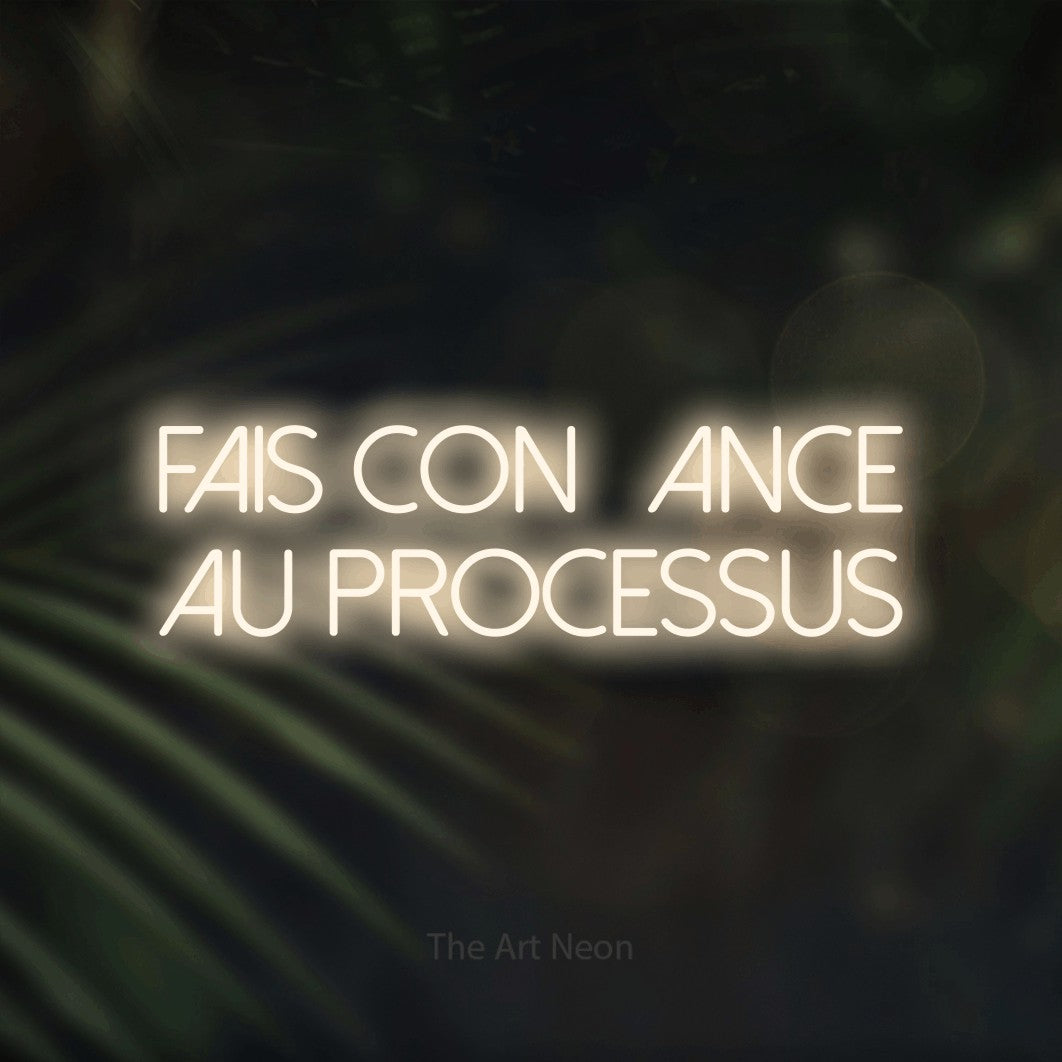 “Fais confiance au processus” LED neon sign in warm white, with thin sans-serif lettering across 2 lines, shown against a blurred palm and bokeh-light backdrop.