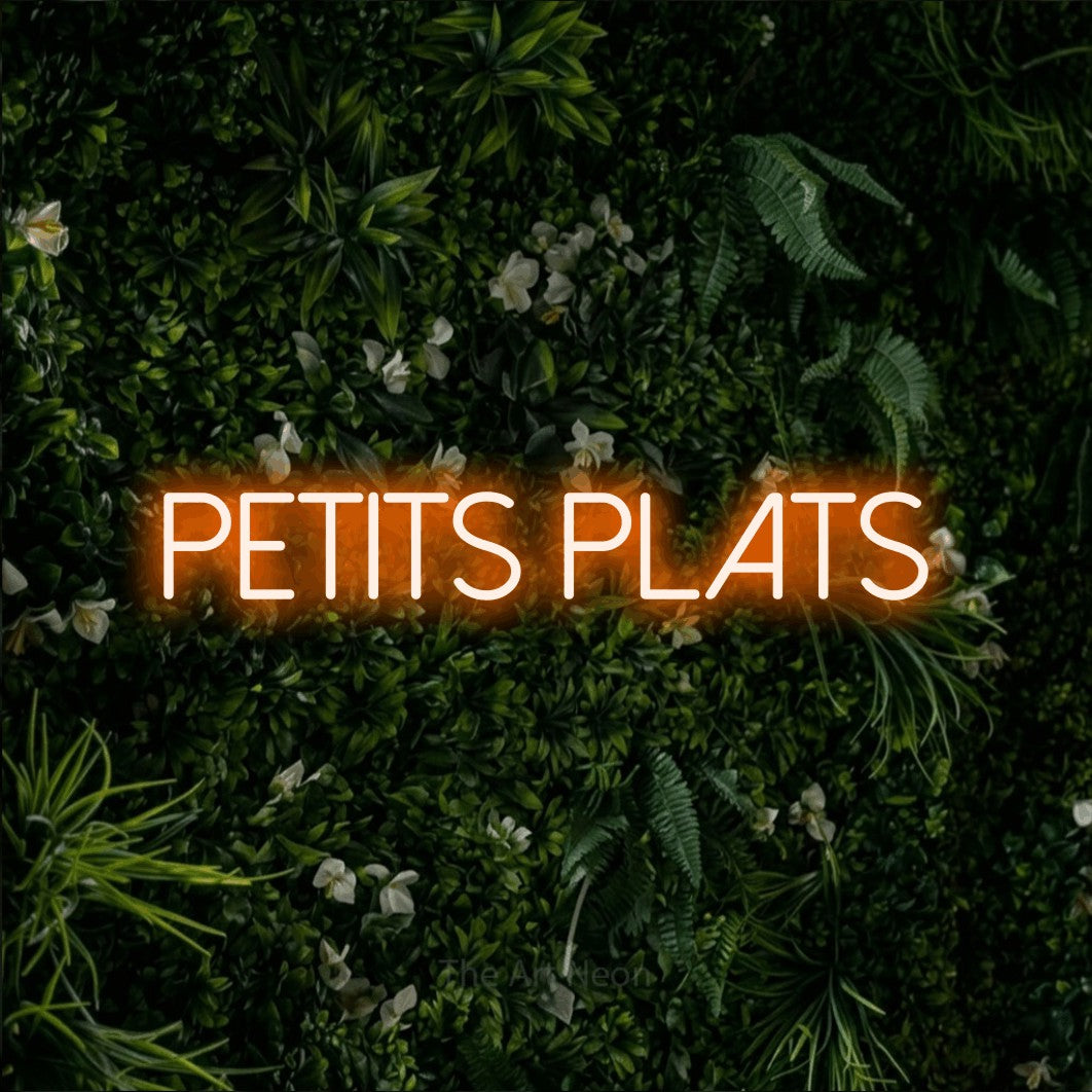 “Petits plats” LED neon sign in orange, with thin sans-serif lettering, shown against a dense green plant wall.
