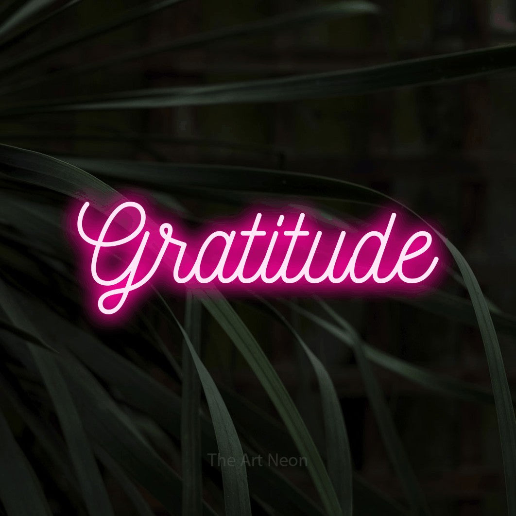 “Gratitude” LED neon sign in hot pink, with ornate cursive lettering, shown against a close-up of dark foliage.