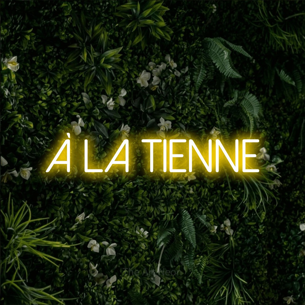 “À la tienne” LED neon sign in yellow, with thin sans-serif lettering, shown against a dense green plant wall.