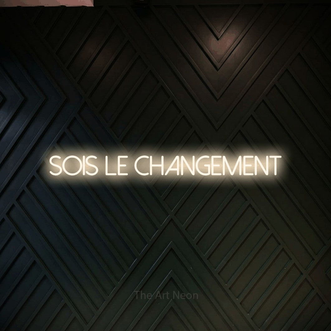 “Sois le changement” LED neon sign in warm white, with thin sans-serif lettering, shown against a black chevron-patterned wood wall.