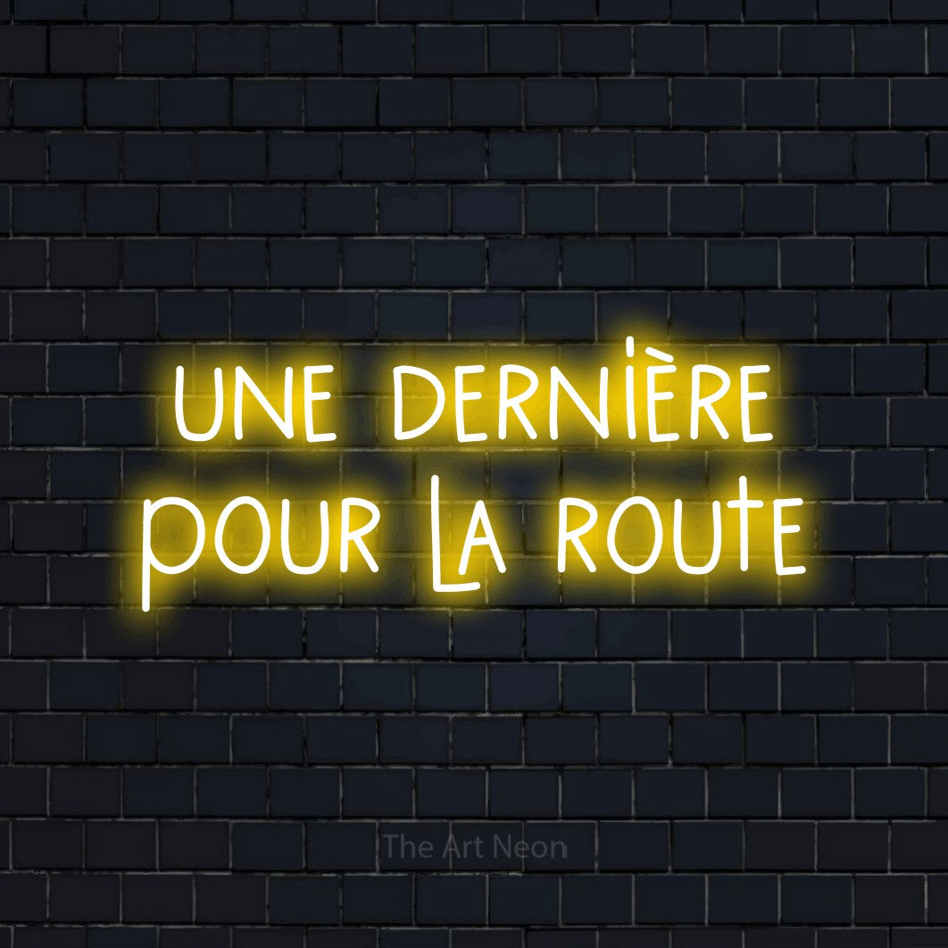 “Une dernière pour la route” LED neon sign in yellow, with narrow hand-drawn lettering across 2 lines, shown against a dark navy brick wall.