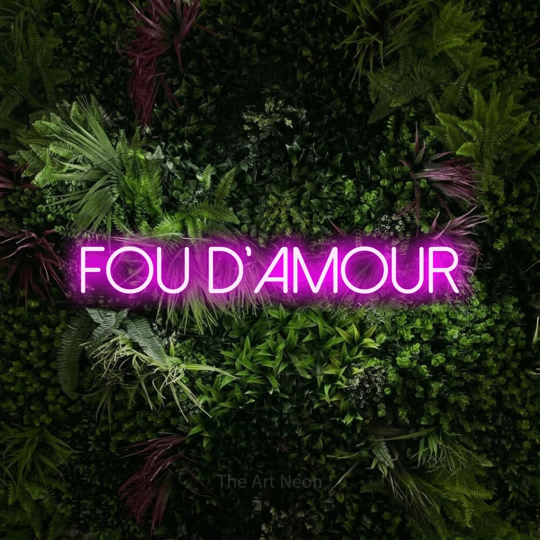“Fou d'amour” LED neon sign in pink, with thin sans-serif lettering, shown against a plant wall with pink flowers.