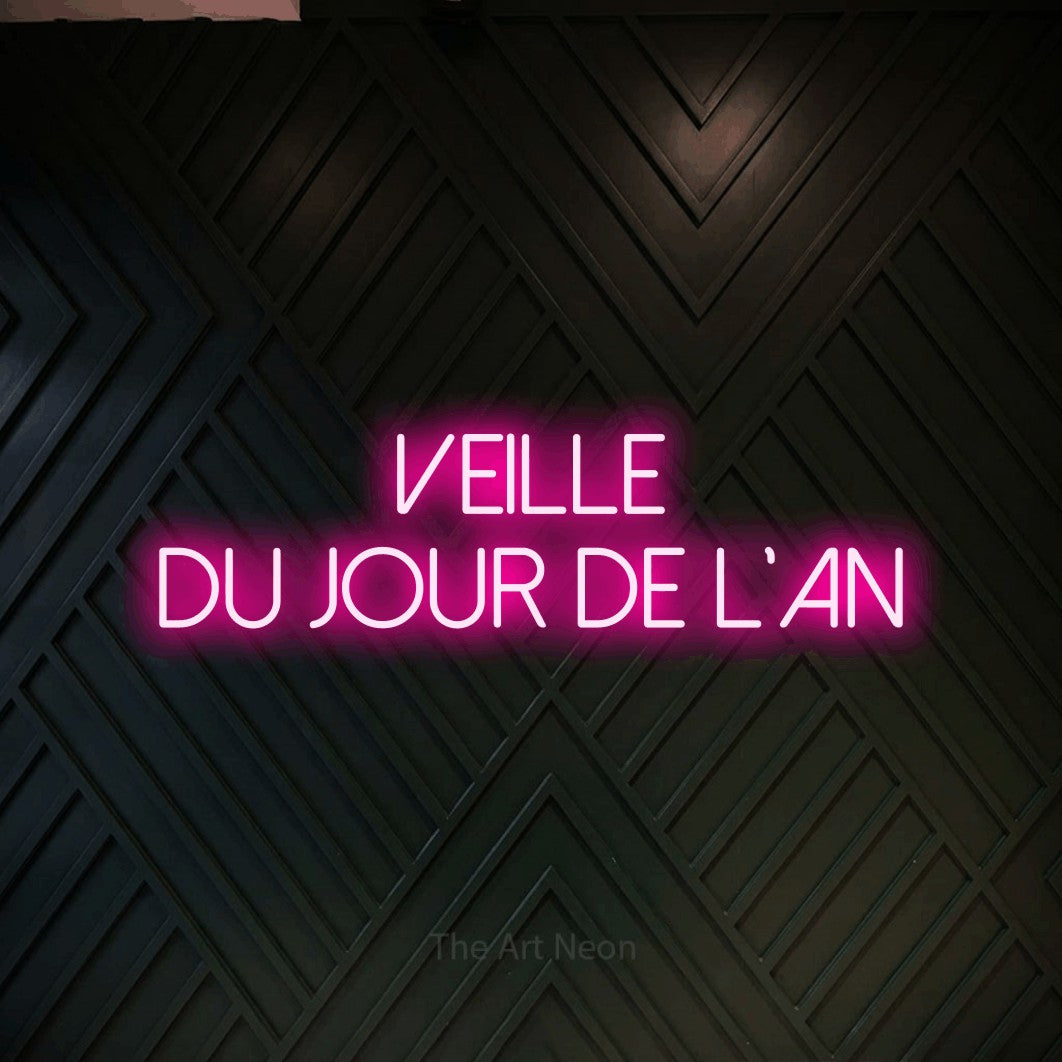 “Veille du jour de l'An” LED neon sign in hot pink, with thin sans-serif lettering across 2 lines, shown against a black chevron-patterned wood wall.