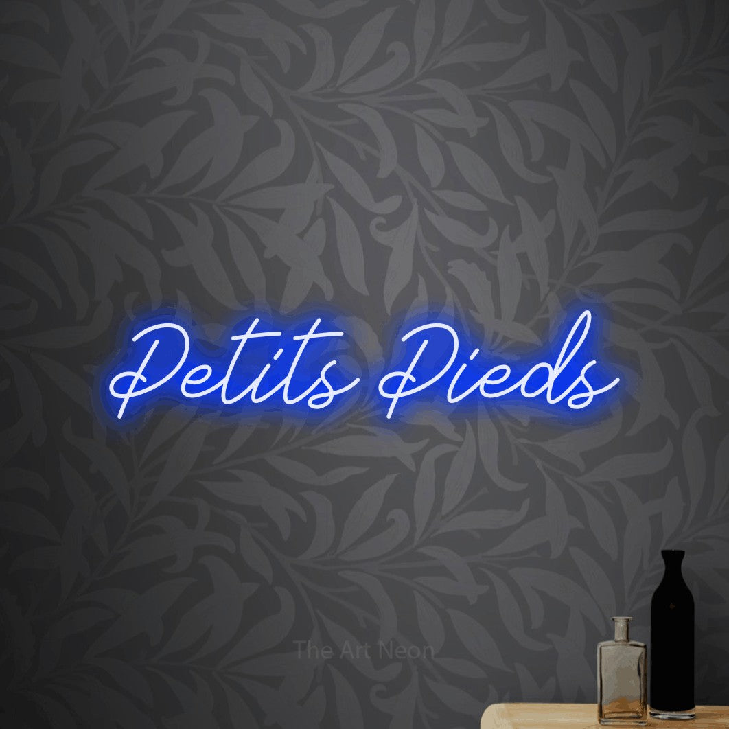 “Petits pieds” LED neon sign in blue, with cursive lettering, shown against grey botanical wallpaper.